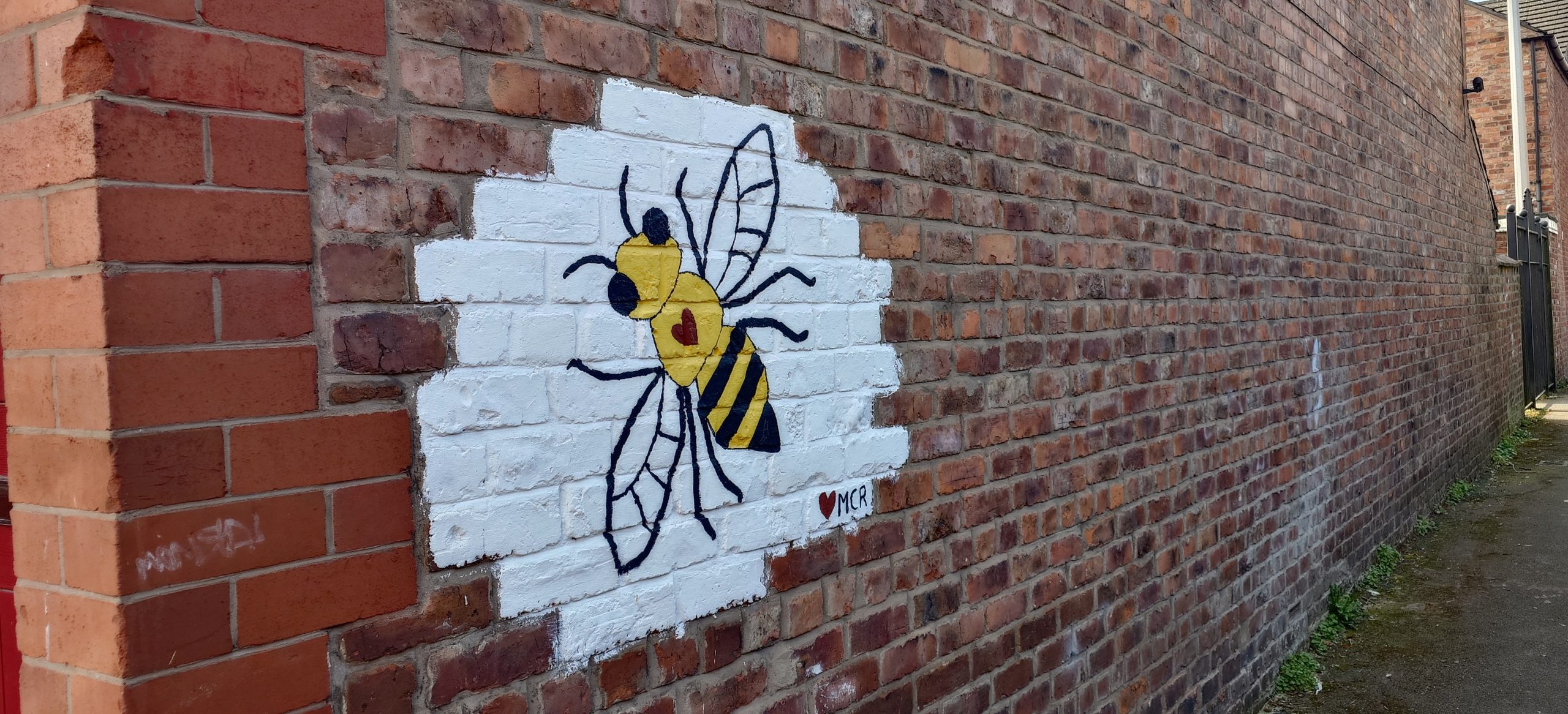A graffiti of a black and yellow bee with a red heart on its side adorns the white patch of a brick wall that stretches along the pathway. Nearby, Nick Midgley's tag "MCR" joins the artwork, making it feel like part of a team creation.