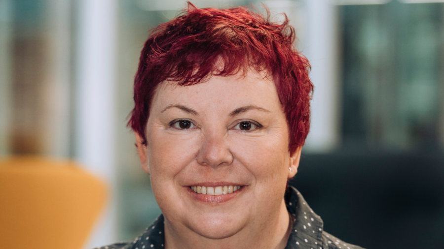 A person with short red hair and a friendly smile is wearing a dark, polka-dotted shirt. The background is softly blurred with tones of white, grey, and yellow.