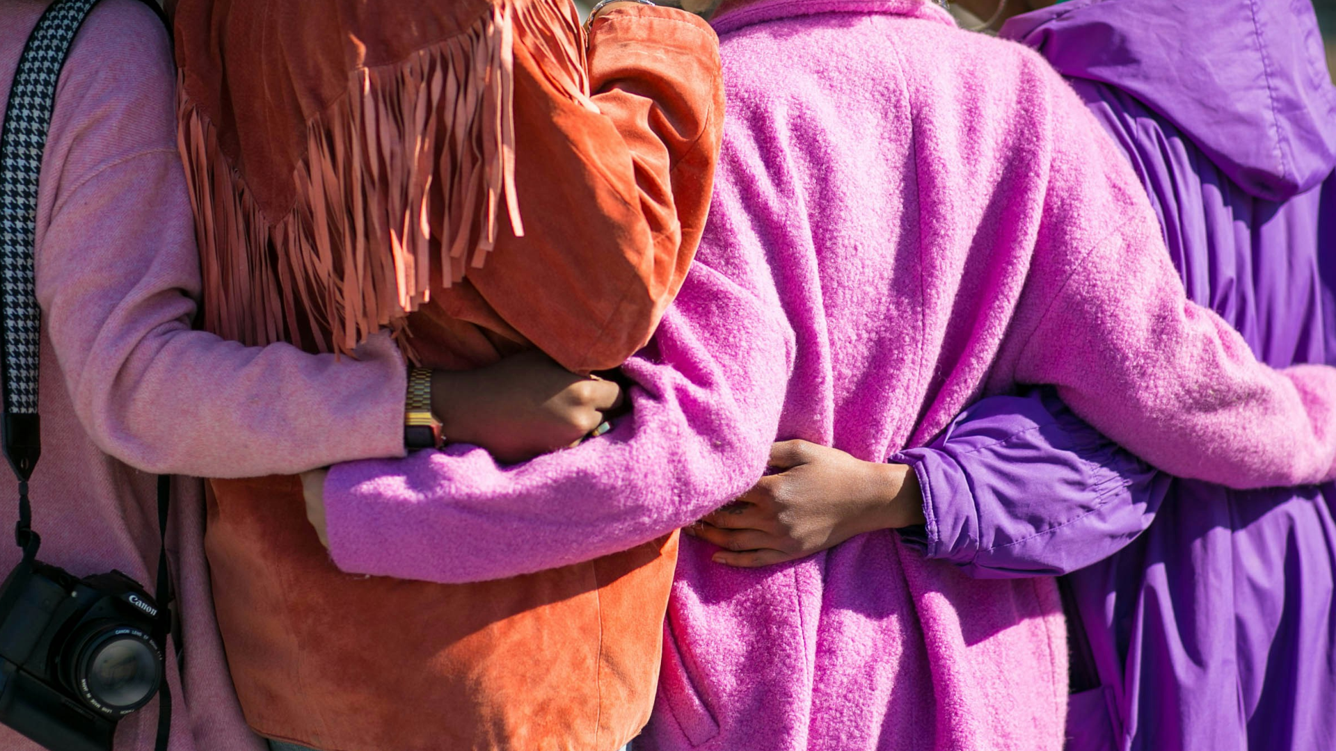 Four people stand side by side with their arms around each other’s backs, wearing vibrant coats in shades of pink, orange, and purple. One person has a camera hanging from their shoulder.