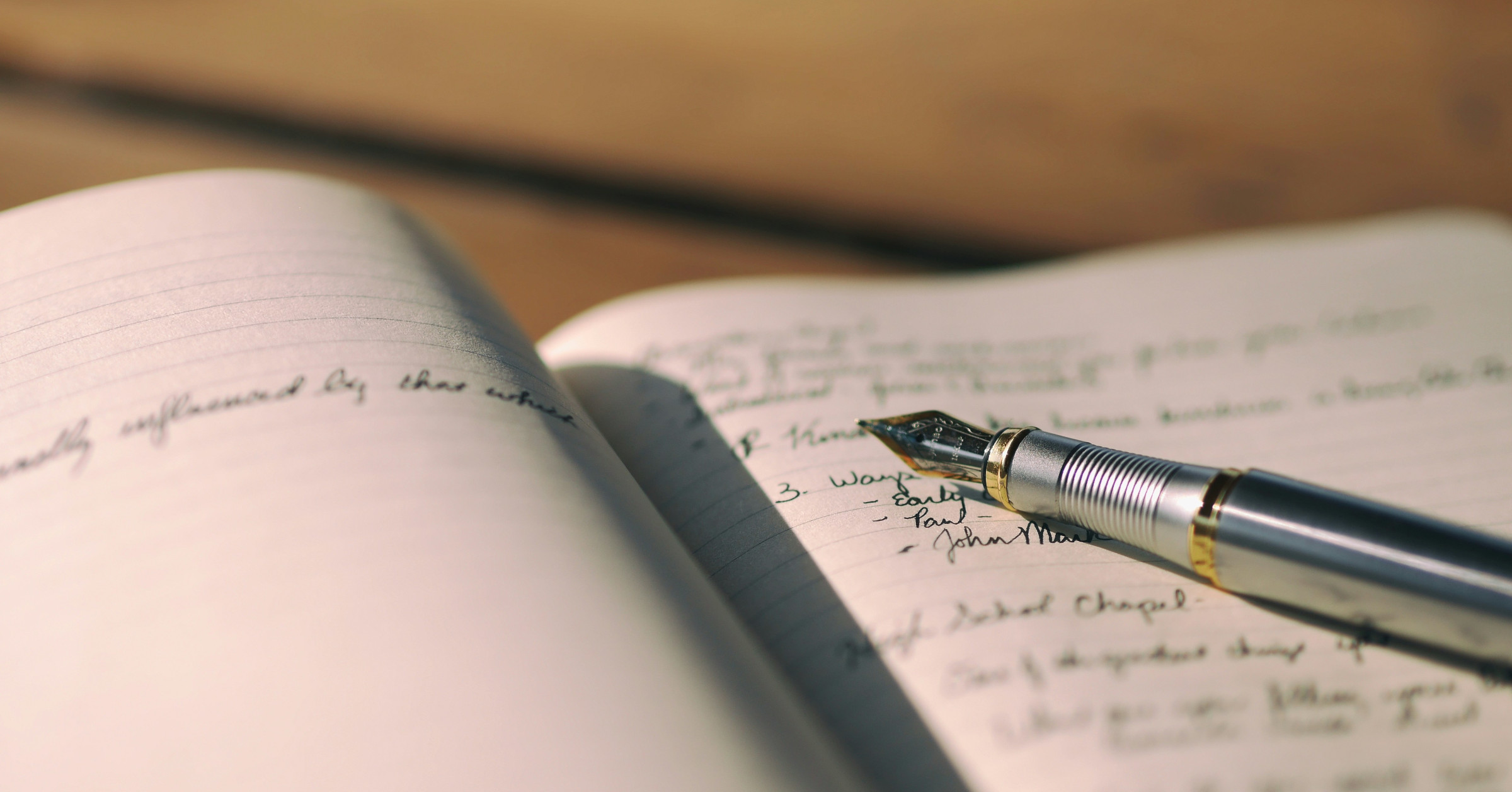 Close-up of an open notebook filled with elegant handwriting in cursive. A fountain pen with a metallic tip rests on the pages, praising the timeless charm of the art of writing. The background is softly blurred, focusing on the pen and notes.