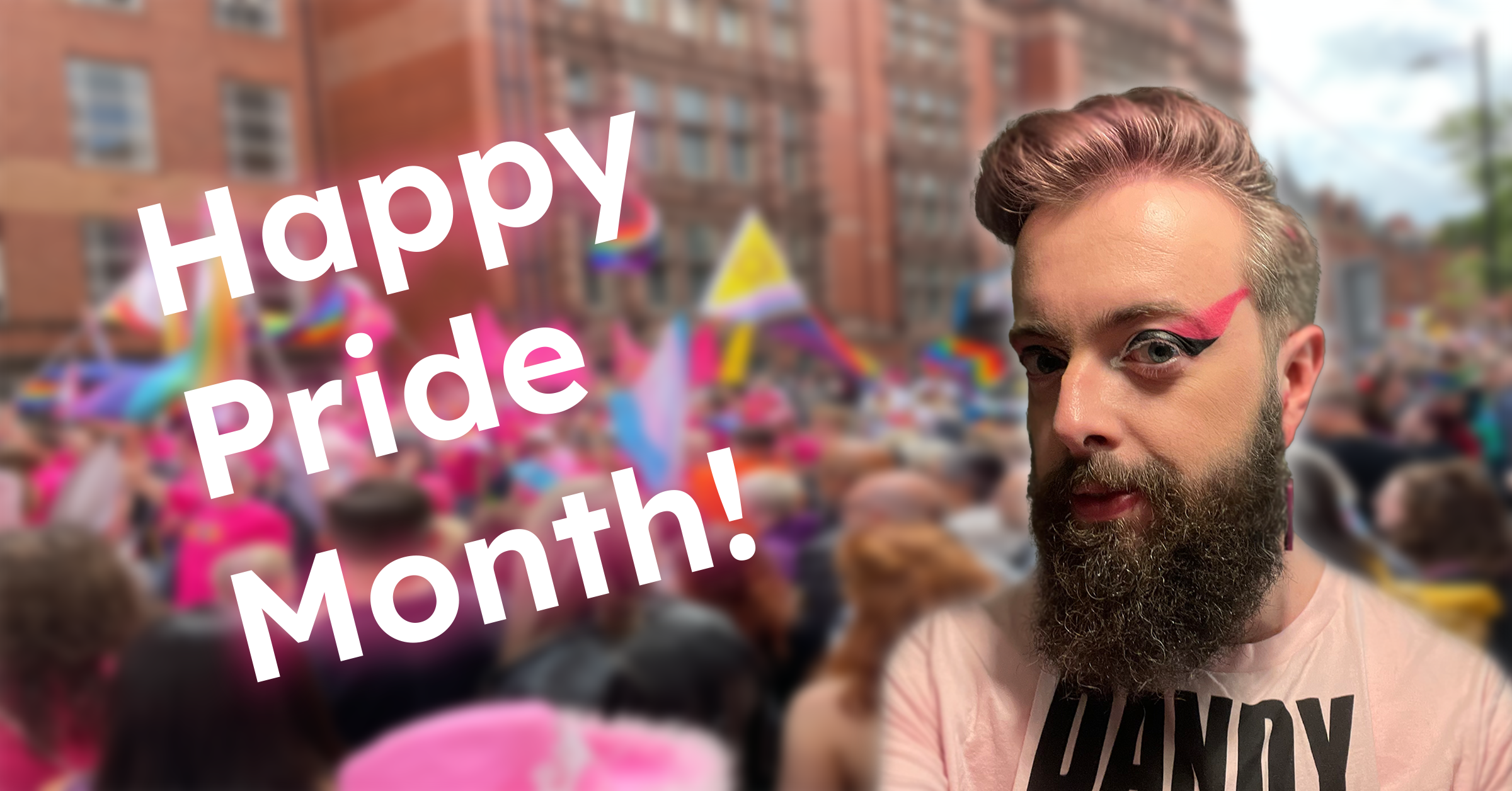 A bearded person with pink makeup and styled hair stands in front of a lively crowd at a Pride parade. Bold text on the image reads, "Happy Pride Month!" Elliot joins the celebration as rainbow flags wave in the background.