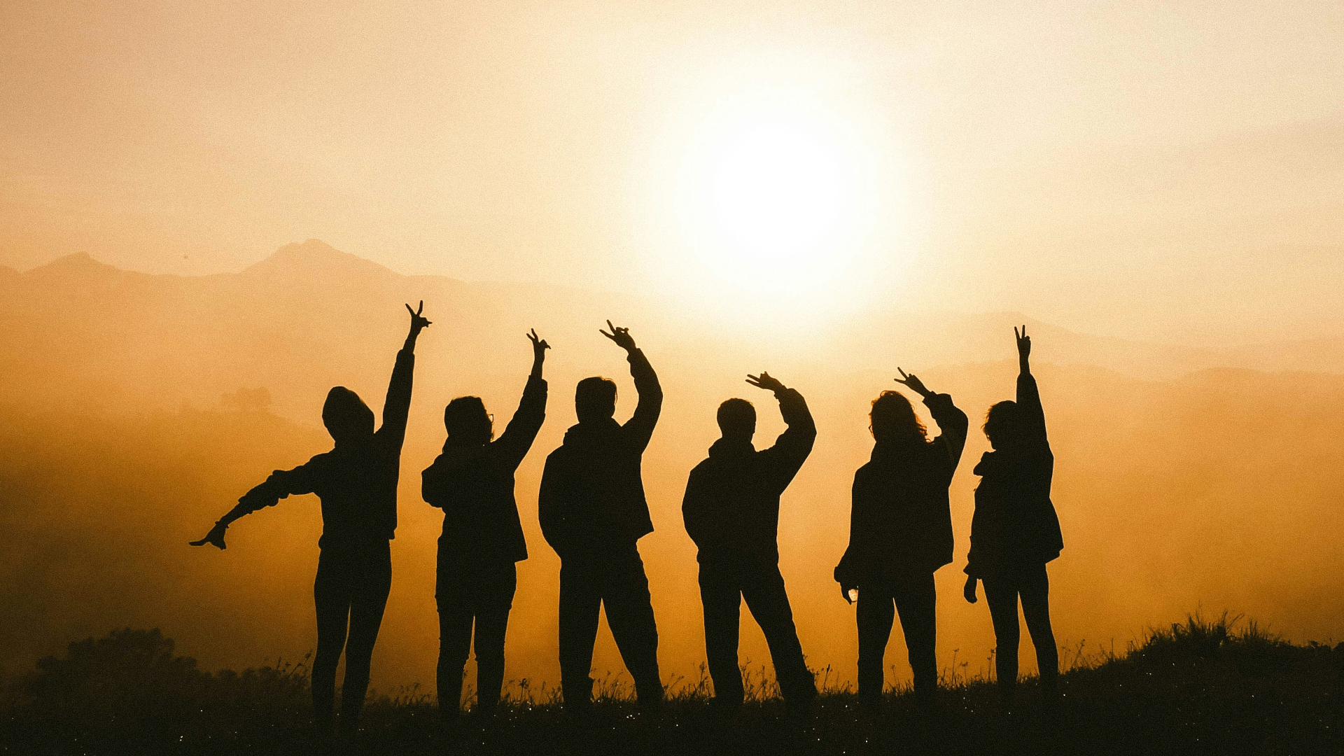 Six people stand side by side on a hill at sunset, silhouetted against the bright sun, with some raising their arms and making peace signs. Mountains and a hazy sky appear in the background.