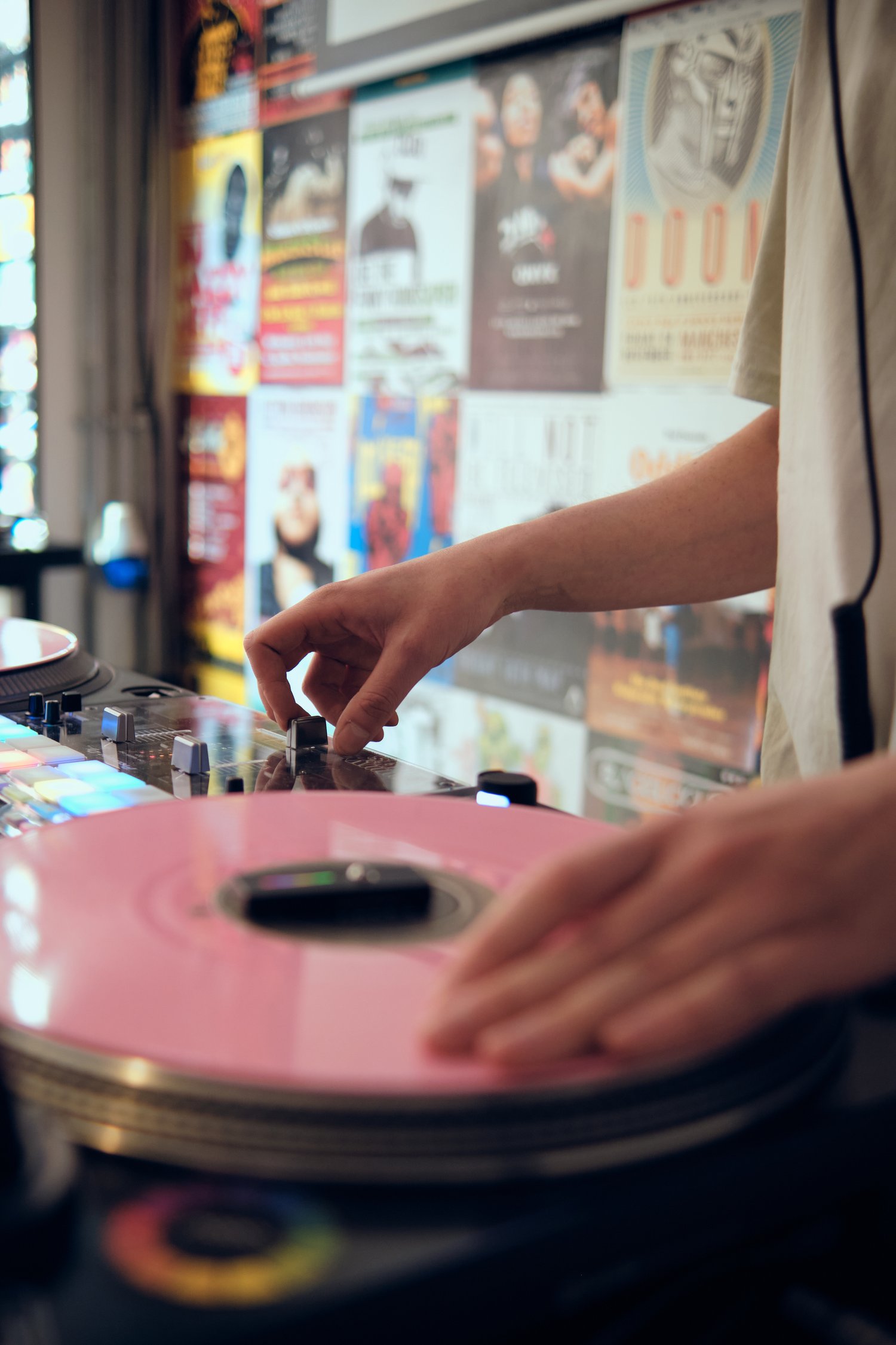 Close-up of a DJ’s hands mixing music on turntables with a pink vinyl record, against a background wall covered with colorful posters.