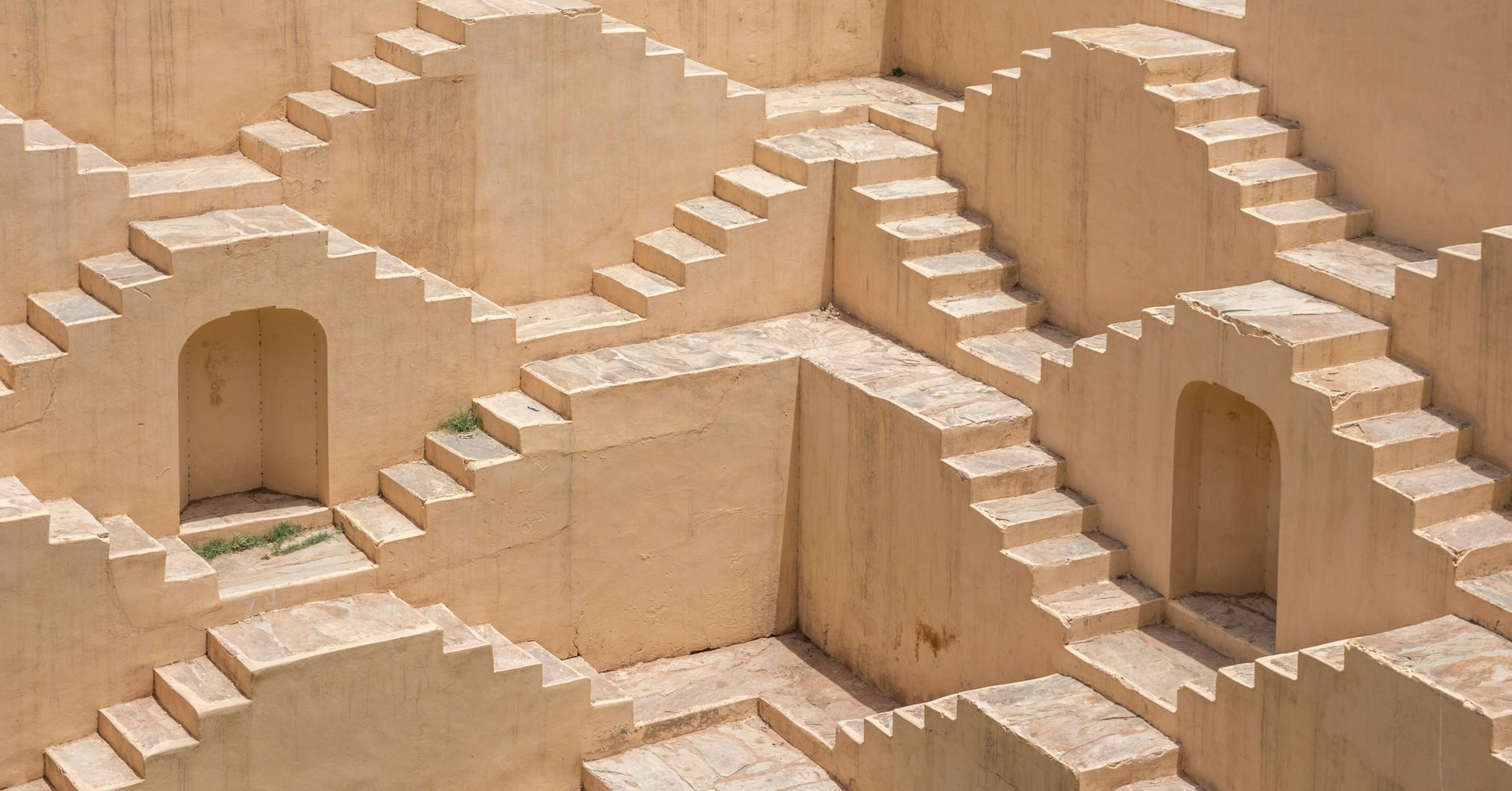 Geometric pattern of pale stone steps and arched doorways form an intricate, maze-like design on a stepped well, where sharp shadows enhance the authenticity and symmetry of the structure’s repeating forms.