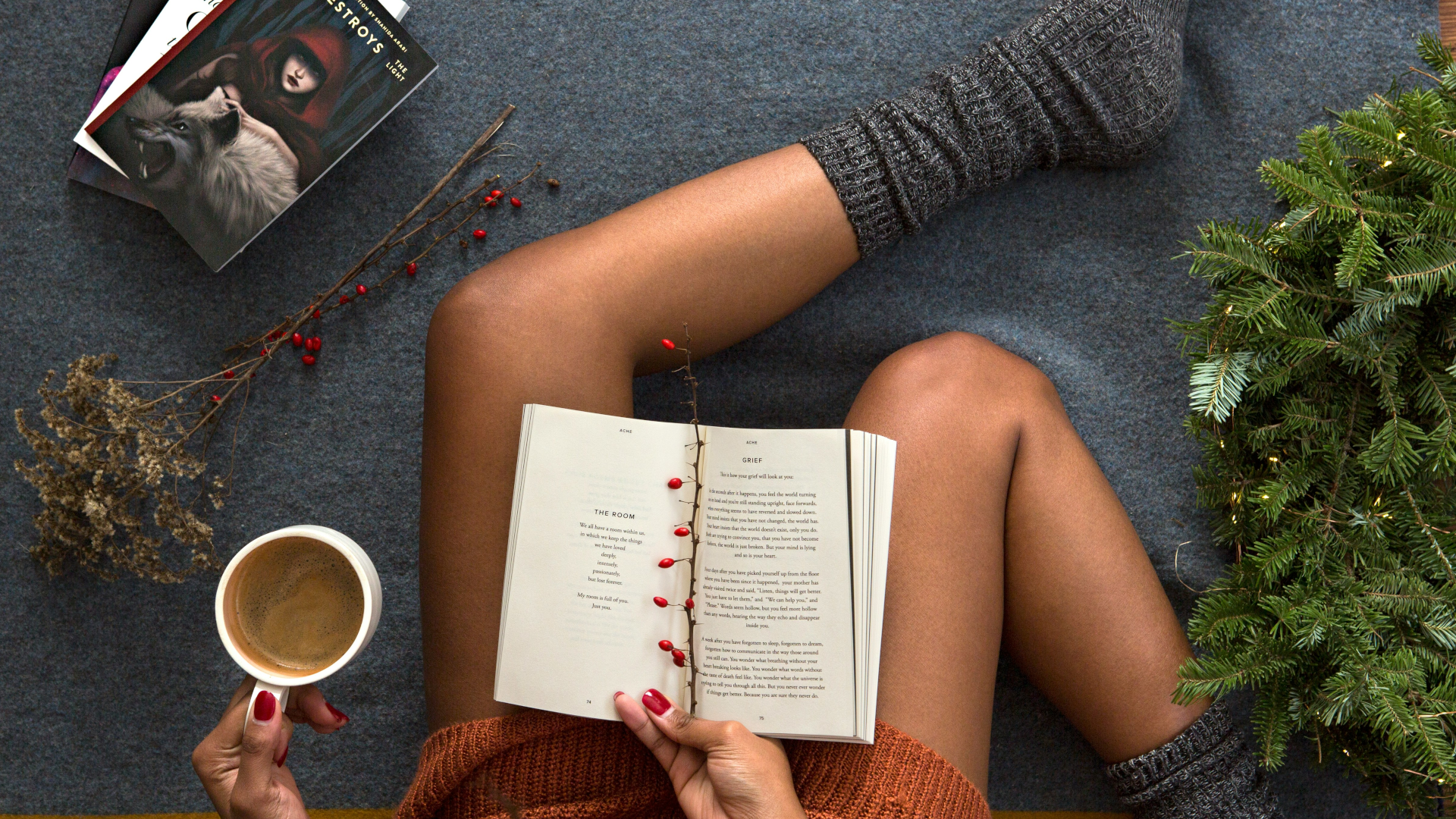 A person with brown skin sits on a gray surface, wearing gray socks and an orange sweater, holding an open book and a cup of coffee, with holiday greenery, berries, and a book on the floor nearby.