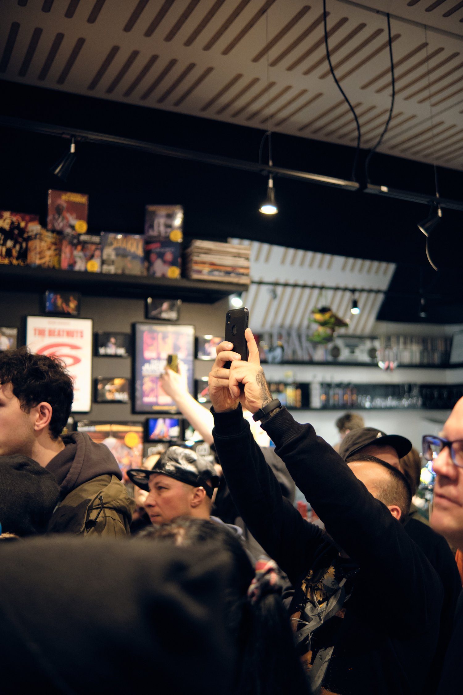 A crowd of people inside a record store, with one person in the center holding up a phone to take a photo or video. Shelves with vinyl records and music memorabilia are visible in the background.